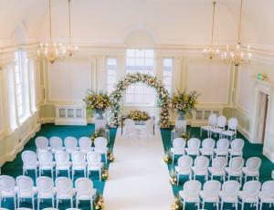 White walled hall with seating arranged for a wedding. Floral arch frames the aisle