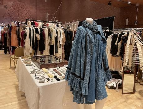 **Alt text:** Vintage clothing and accessories displayed at a fair, with a mannequin wearing a blue dress in the foreground.